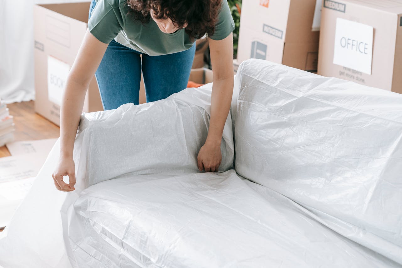 cta-bg A woman carefully wraps a sofa in protective material while packing boxes in a bright room.