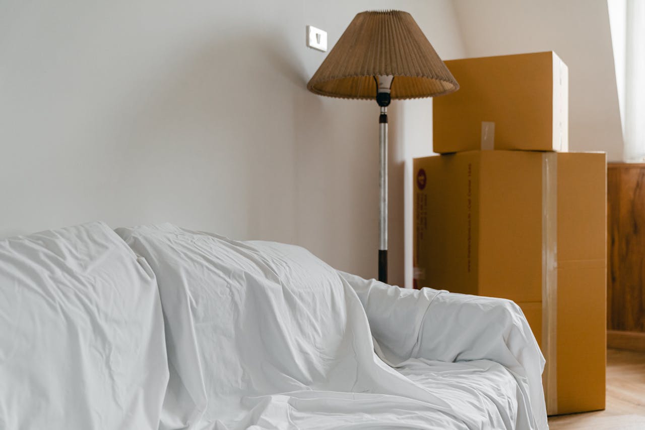 home-hero-bg Covered with sheet sofa and floor lamp and packed cardboard boxes in light spacious room of new contemporary house